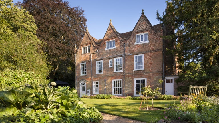 A view of a grand red-brick mansion with a lawn in front and borders and trees to the left, and blue skies above